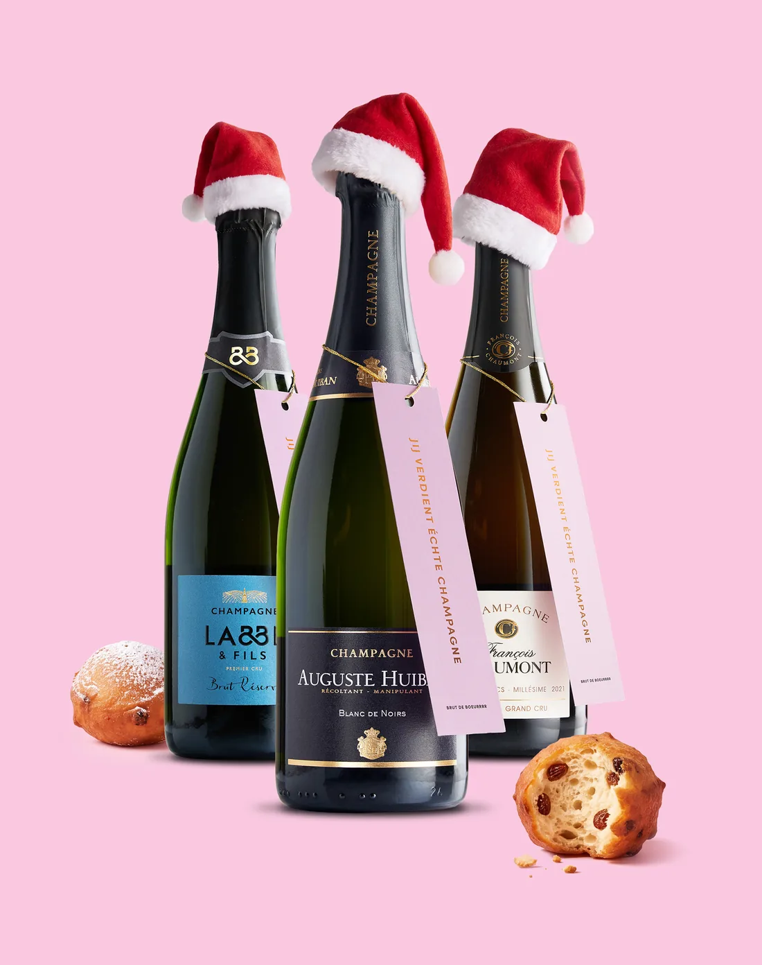 Drie flessen Brut de Boeurrrr-champagne met kerstmutsen en roze halslabels op een roze achtergrond